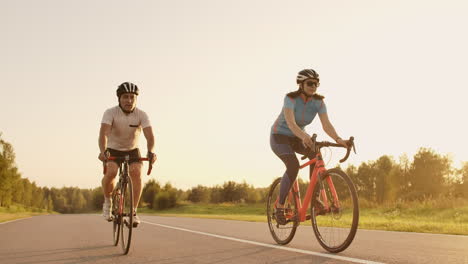 Un-Hombre-Y-Una-Mujer-Montan-Bicicletas-Deportivas-En-La-Carretera-Al-Atardecer-Con-Equipo-Y-Cascos-Protectores-En-Cámara-Lenta-A-120-Fps.